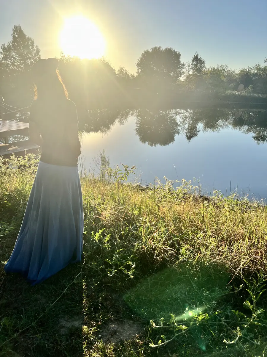 A woman in a long blue skirt watching the sun rise over the pond